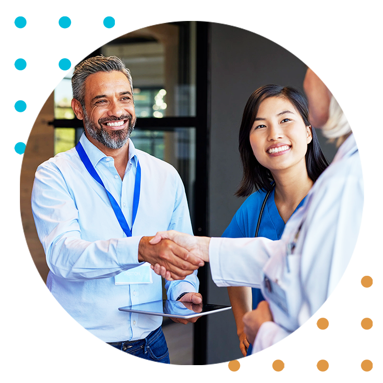 Man in a blue button down shirt holding an ipad is shaking hands with a woman in a tab coat. Another woman in blue scrubs is smiling in the background.