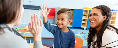 school psychologist giving a young boy with a blue shirt a high five in a colorful classroom