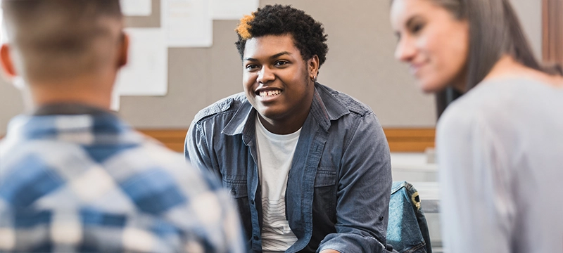 A high school boy smiles at another student facing him in a circle of students meeting, representing how the BRIEF2 helps students with special needs find success