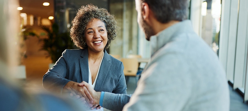 A woman in professional attire smiles and shakes hands with a man in a modern office setting