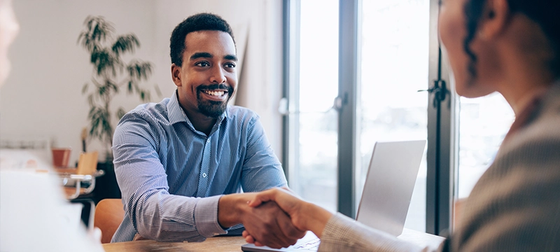 A man smiles while shaking a woman's hand at a desk, representing him getting hired after taking the SDS and finding the right career