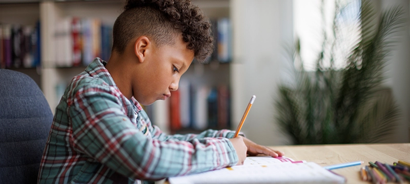 A young boy looks down at his school workbook while writing in it with a pencil