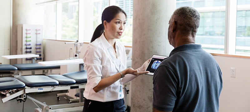 A woman working in a healthcare setting holds a tablet to show and explain a brain scan to a male patient, representing Brain Injury Awareness Month and discussing symptoms