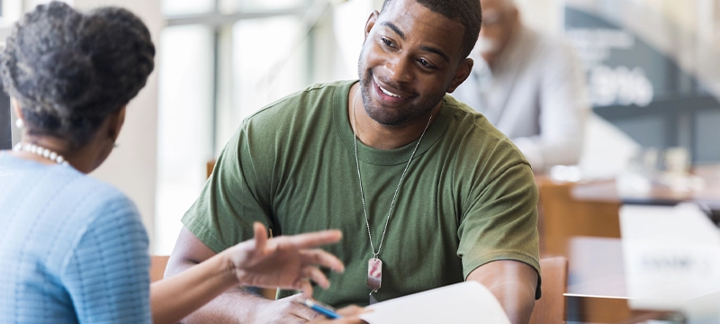 A military veteran wearing a green shirt and dog tag necklace sits across from an older woman smiling and discussing his resume to convey how taking the SDS can help veterans to find a career