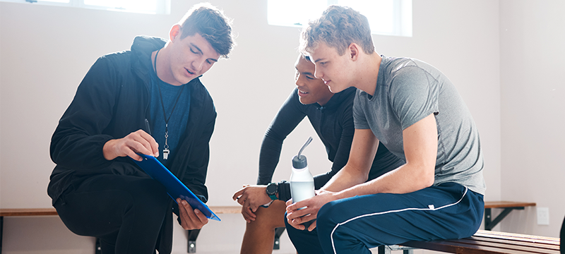 A coach kneels in front of two players seated on a bench while explaining to them a strategy from a clipboard