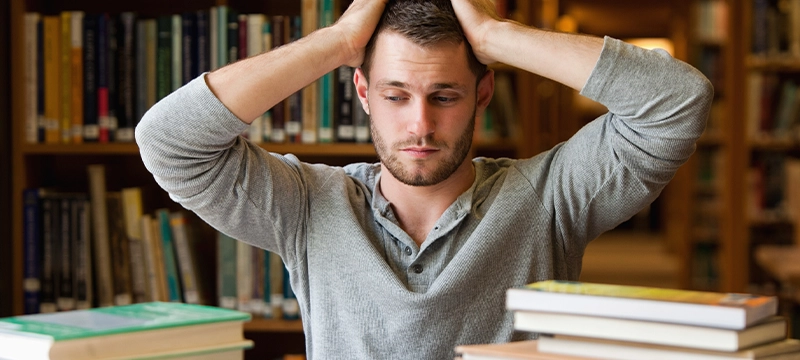 Stressed adult man with hands on his head, sitting at a desk with books, representing the executive function challenges assessed by the BRIEF2A tool for adult ADHD