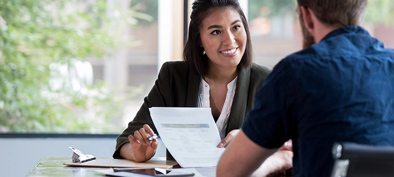 A woman smiles while showing a slip of paper to the man sitting across from her, representative of a psychologist discussing insurance billing to a patient or fellow psychologist